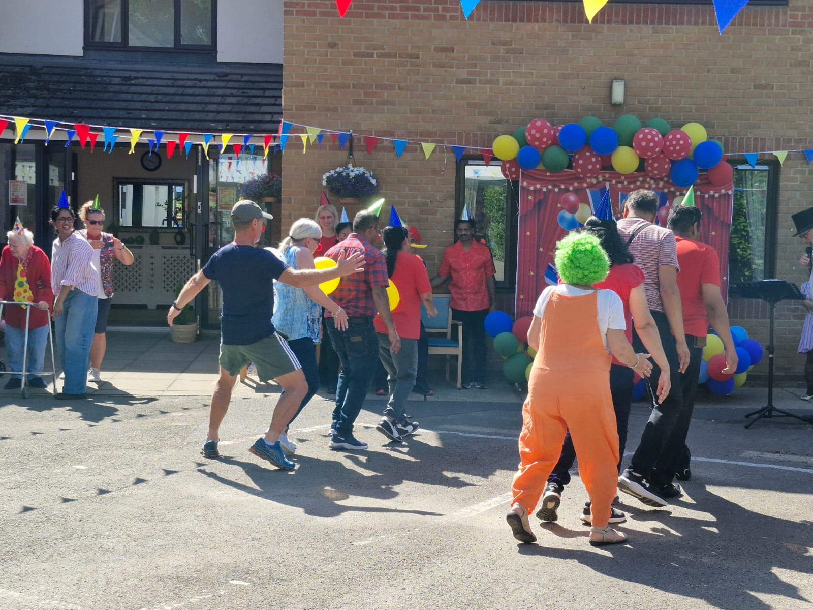 Residents and staff engage in a lively dance outside, surrounded by colourful decorations, balloons, and bunting for a festive event.