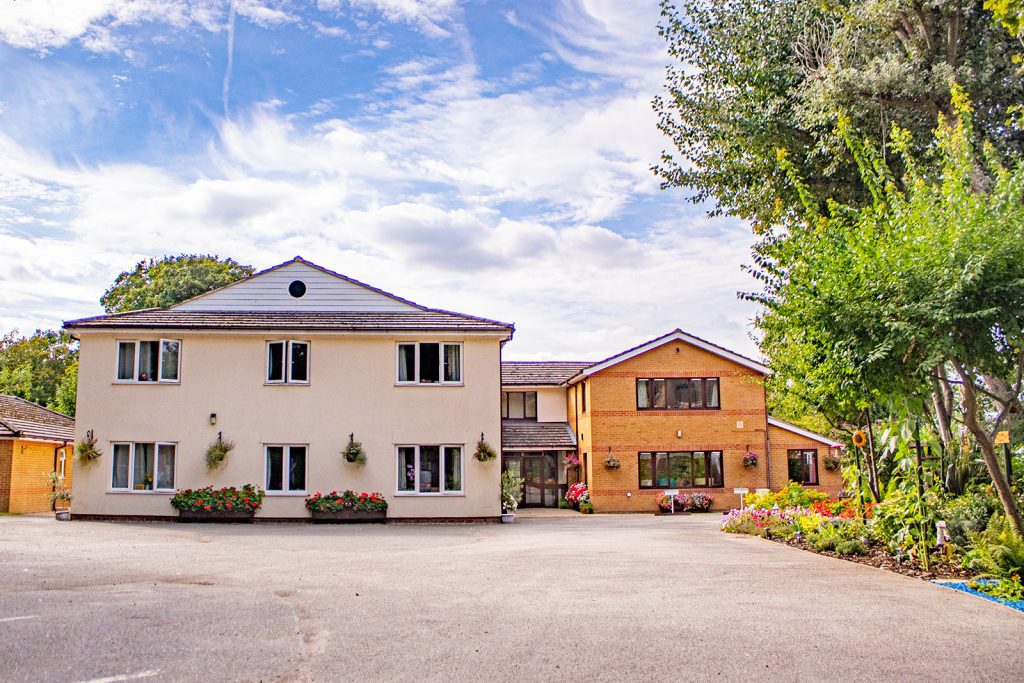 Two-storey care home with a welcoming facade, surrounded by landscaped gardens, bright flowers, and a clear blue sky above.