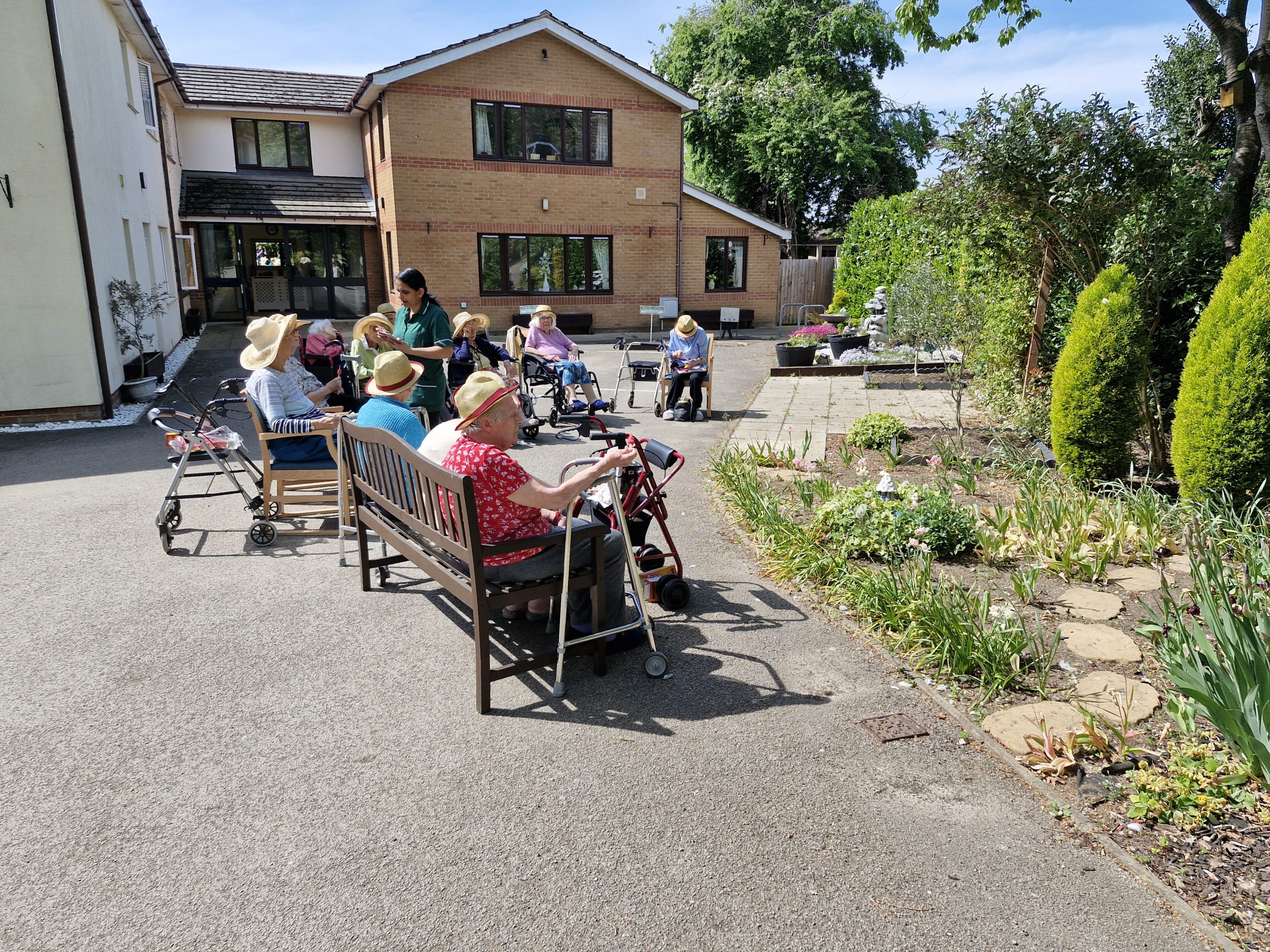 Residents sit on benches outside Stanway Green Lodge, enjoying the sun near a garden, with staff assisting and greenery in the background.