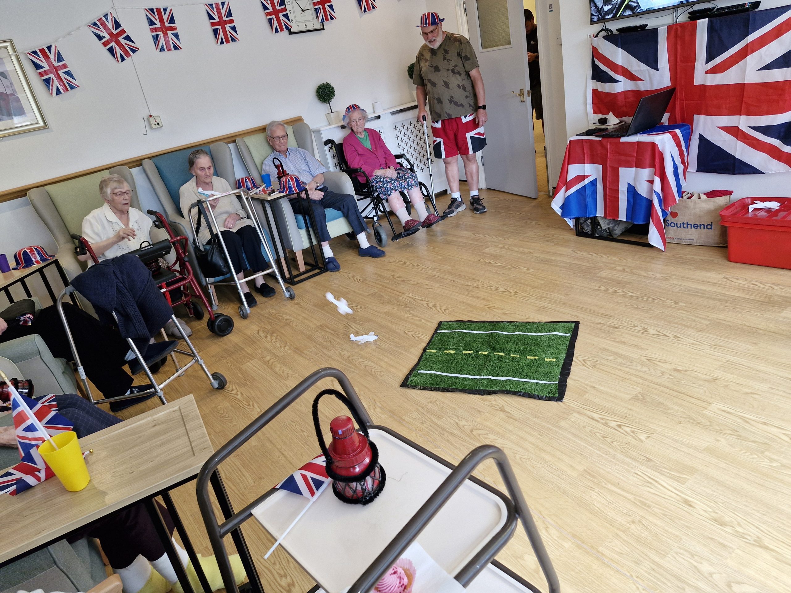 Residents in a lounge decorated with Union Jack flags engage in activities. A green mat with a marked path is visible, alongside supportive seating.