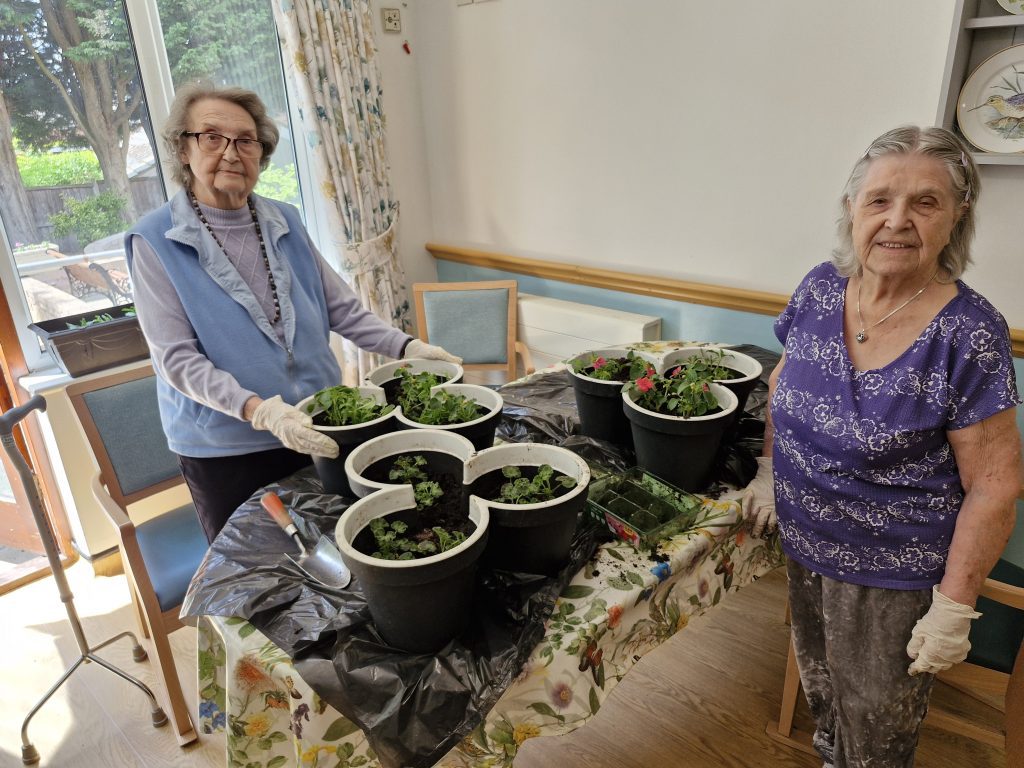 Two elderly women, gloved and smiling, stand by a table filled with potted plants, surrounded by a bright, cheerful indoor space.