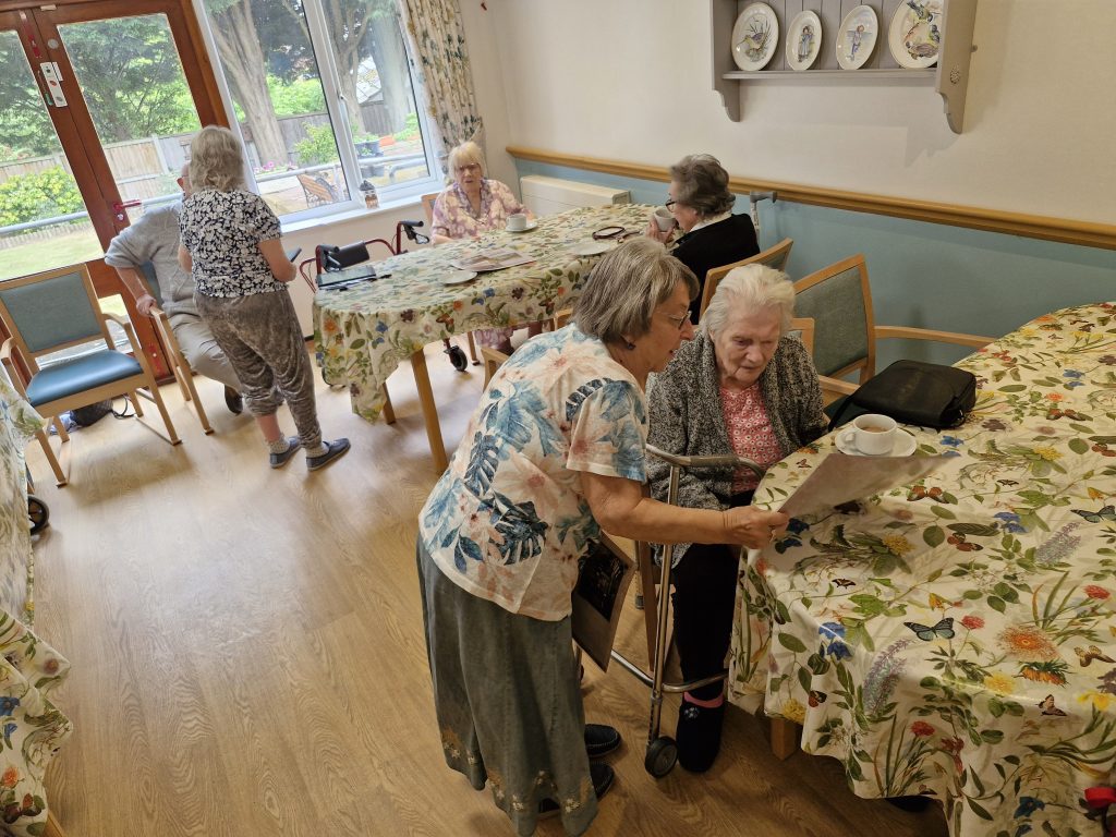 A care home activity room with floral tablecloths, where staff assist residents with tea and conversation amidst a bright, welcoming atmosphere.