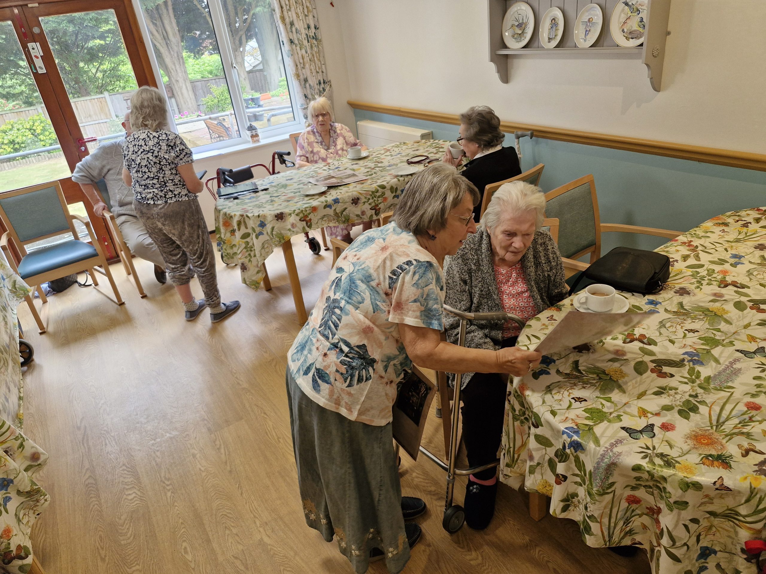 A care home activity room with floral tablecloths, where staff assist residents with tea and conversation amidst a bright, welcoming atmosphere.