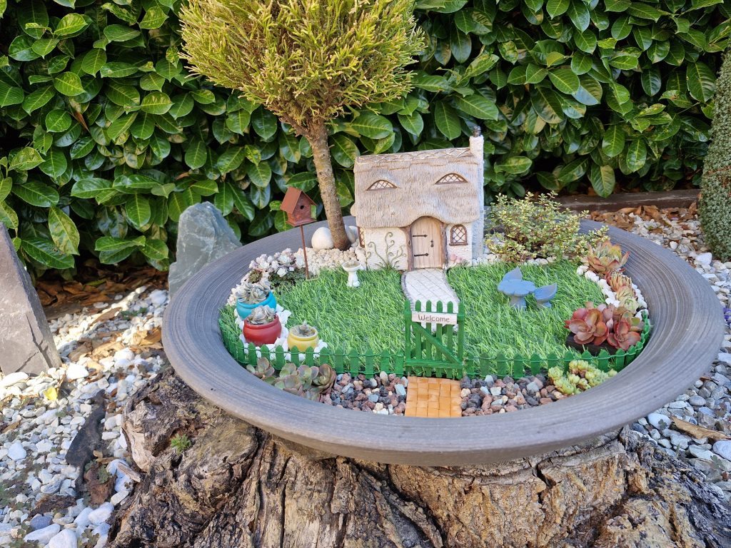 A charming miniature garden scene featuring a thatched cottage, decorative plants, a small tree, and a picket fence, surrounded by lush greenery.