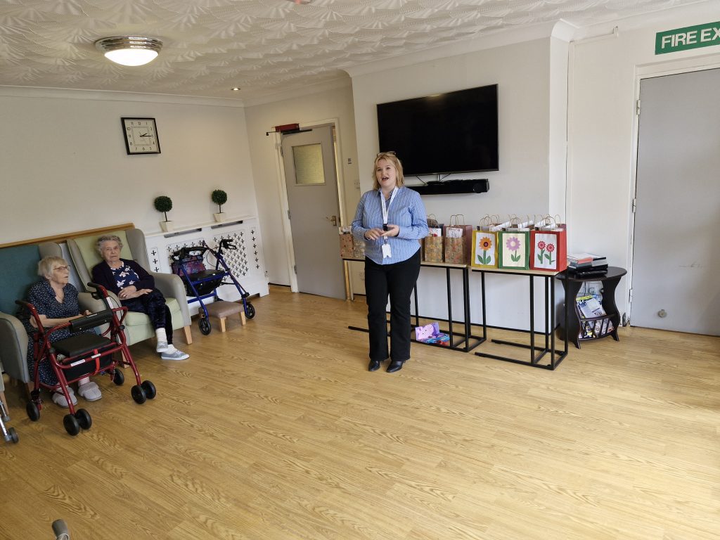 A staff member presenting to residents in a bright, spacious lounge with wooden floors, two elderly women seated, and decorative boxes displayed.