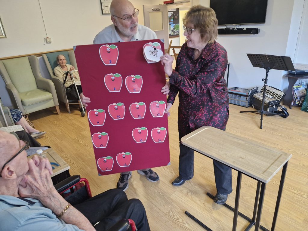 Two staff members engage residents in a care home activity, showcasing a large board with apple illustrations, as residents watch.