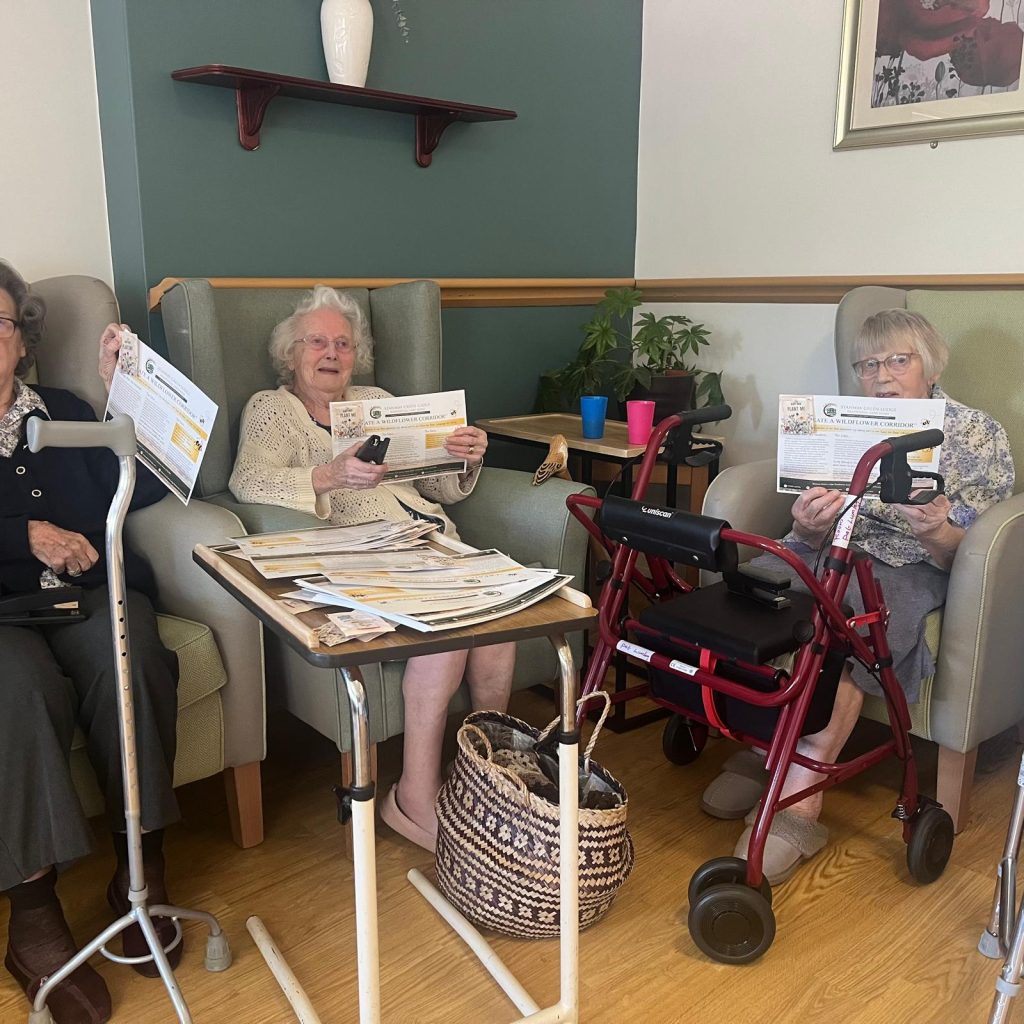Three elderly women in armchairs hold newsletters and smile, surrounded by a warm, inviting lounge with plants and a table.