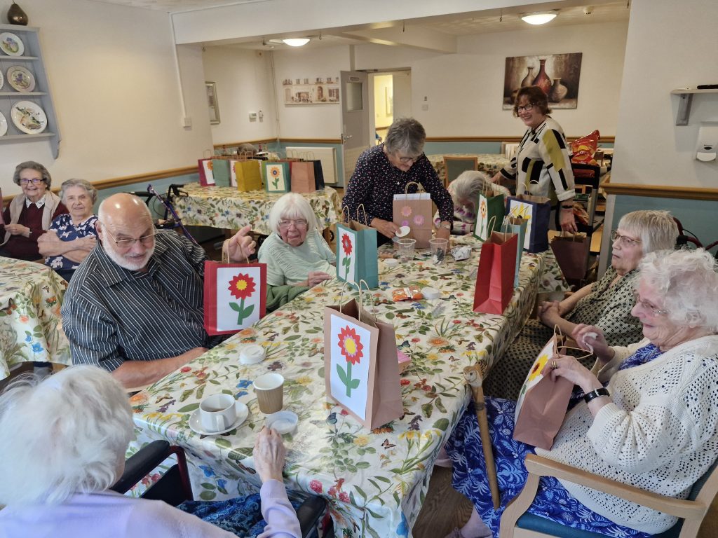 Residents enjoying a social gathering at Stanway Green Lodge, with floral-themed bags on tables and cheerful decorations in a warm, inviting space.