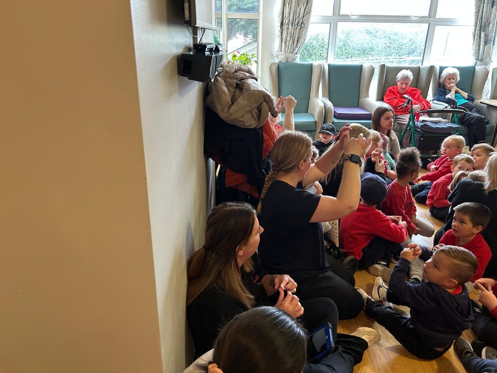 A warm gathering in a care home lounge, with staff and children in red clothing interacting, surrounded by seated elderly residents.