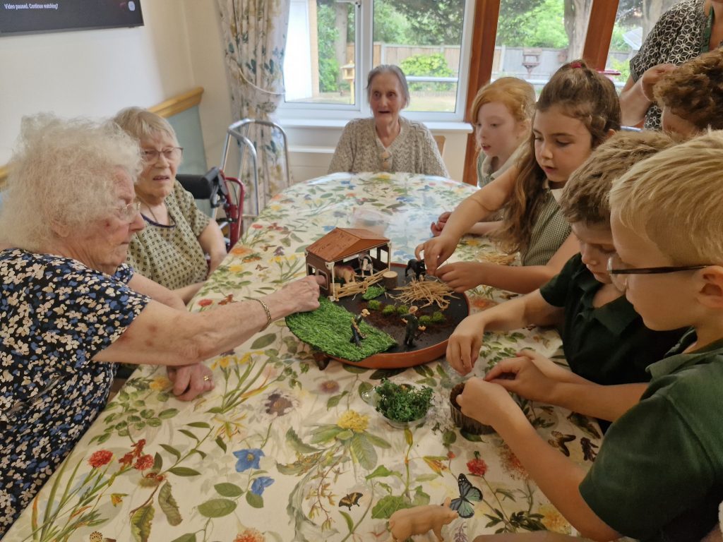 Elderly residents engage with children around a table, crafting a model garden, surrounded by a floral tablecloth and bright natural light.
