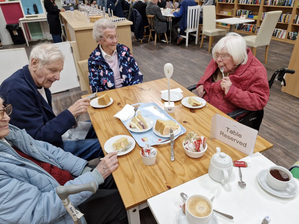 Residents enjoying tea and cake at a wooden table in a warm, inviting café setting, with reserved signage and a friendly atmosphere.