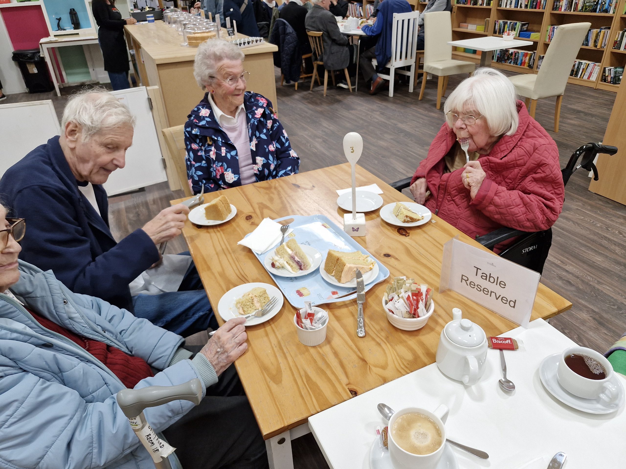 Residents enjoying tea and cake at a wooden table in a warm, inviting café setting, with reserved signage and a friendly atmosphere.