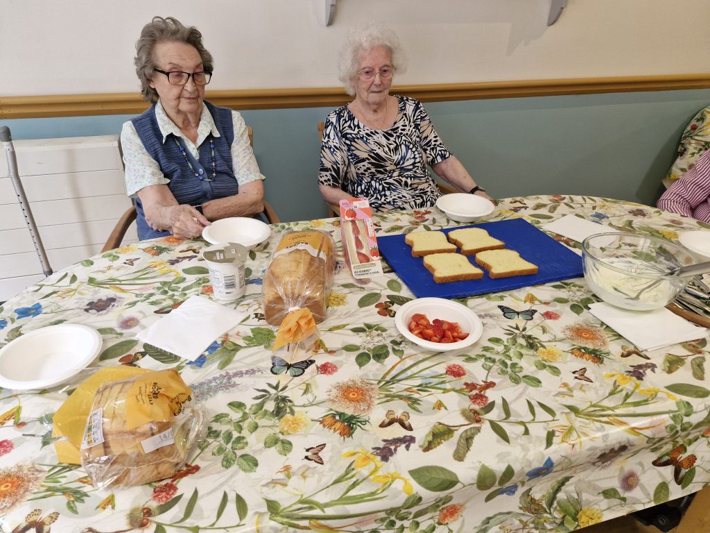 Two elderly women sit at a table covered with a floral tablecloth, with bread, tomatoes, and a bowl of cream on display for preparing sandwiches.