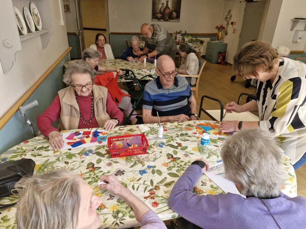 Residents engage in arts and crafts at a table covered with a floral tablecloth, with vibrant materials and a staff member assisting.