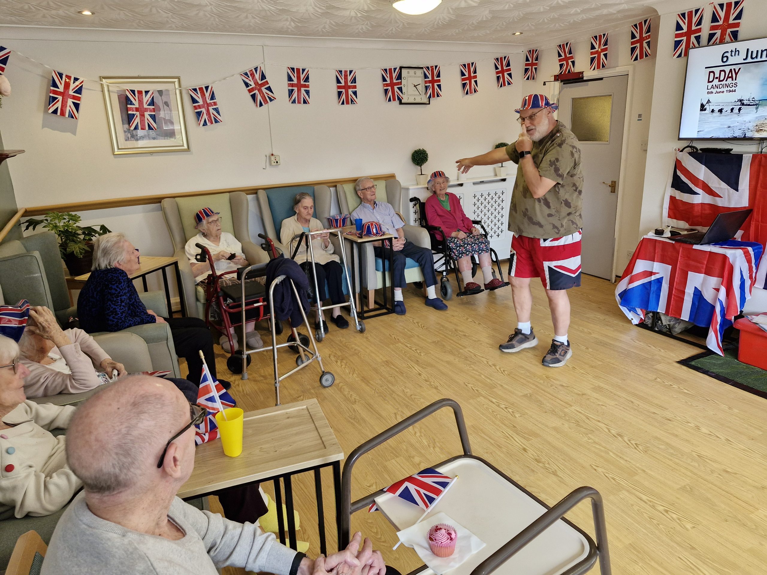 A lively room decorated with Union Jack flags, featuring residents engaged in an event while one man gives a speech, all wearing festive hats.