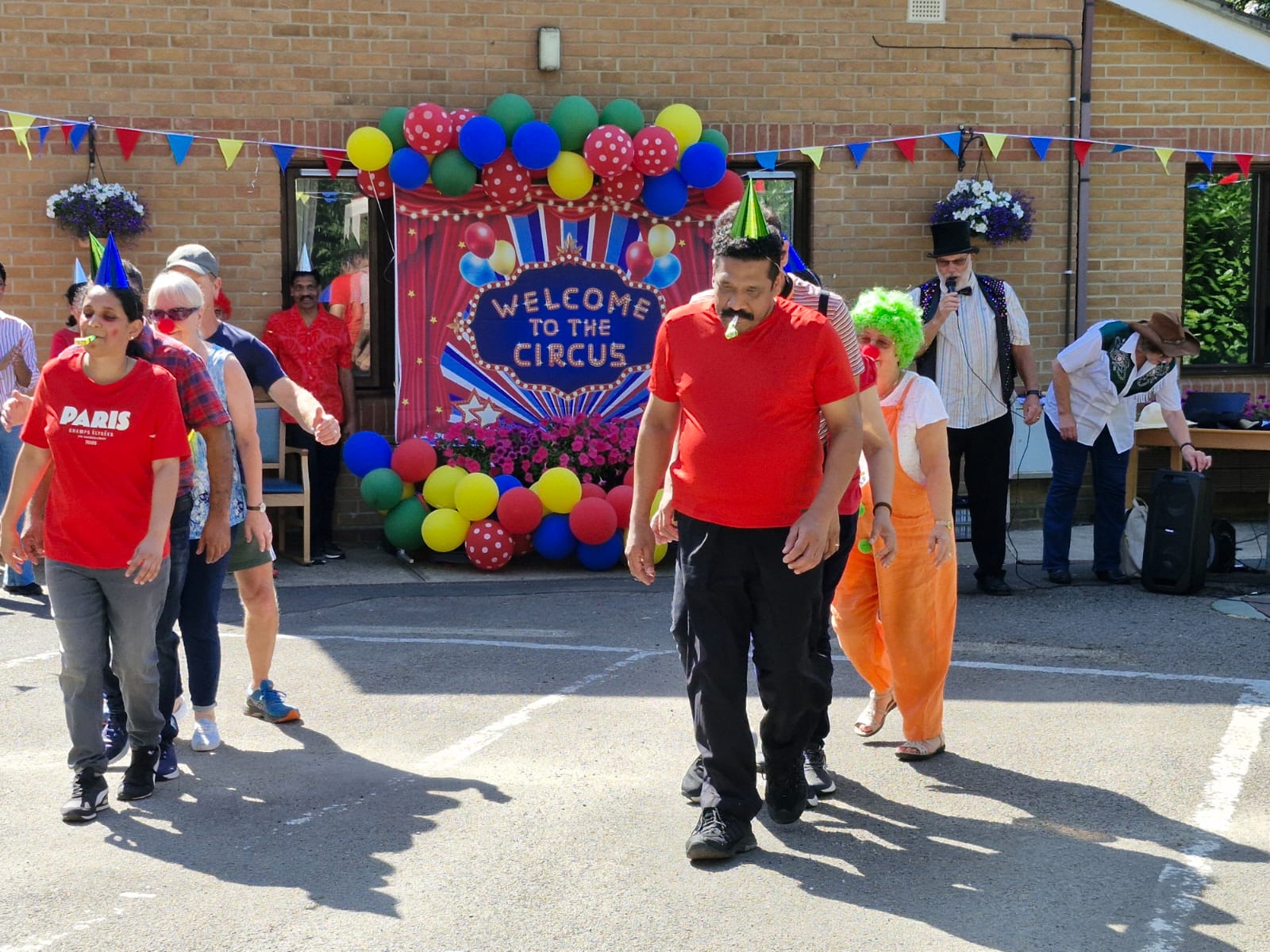 Residents and staff participate in a lively circus-themed event, with colourful balloons, decorations, and playful outfits in a sunny outdoor setting.