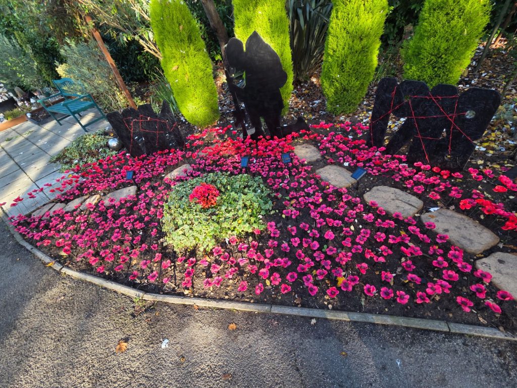 A colourful flower bed features red and pink blooms surrounding a circular green arrangement, framed by dark stone figures and greenery.