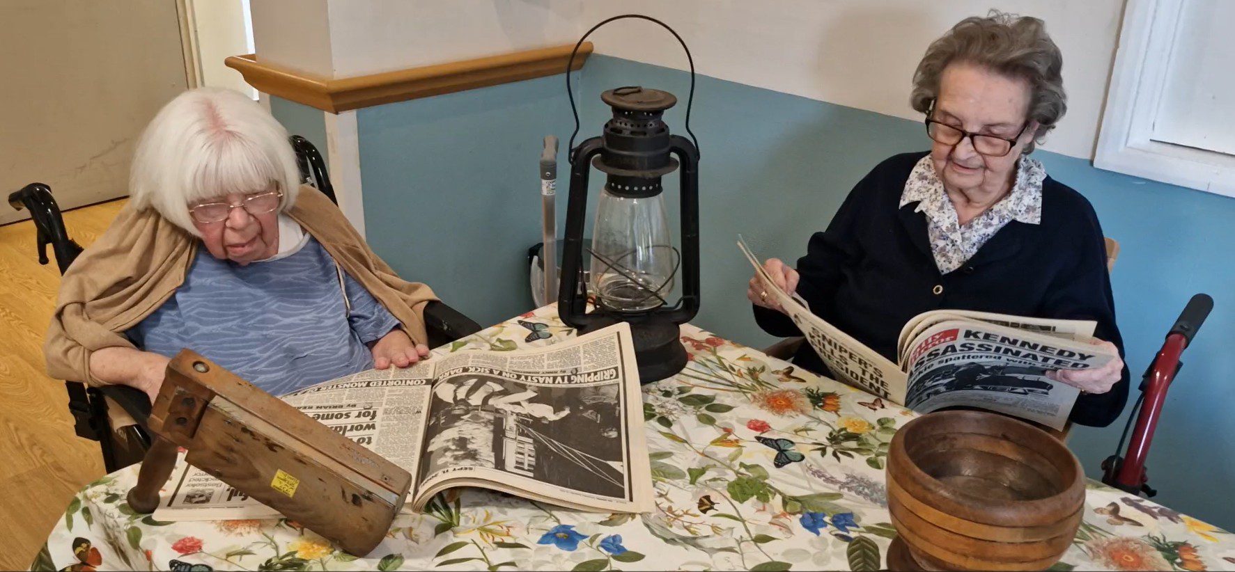 Two elderly women sit at a table covered with a floral tablecloth. One reads a newspaper, while the other examines an old wooden object beside an antique lantern.