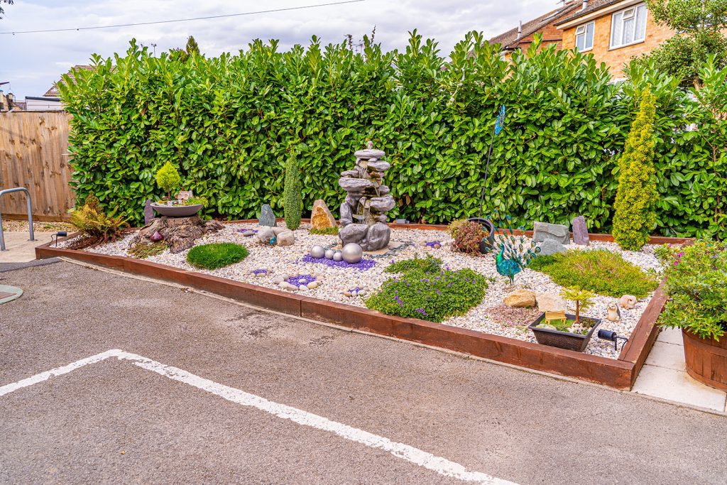 A tranquil garden features a stone water fountain surrounded by decorative stones, greenery, and a variety of plants in organised beds.