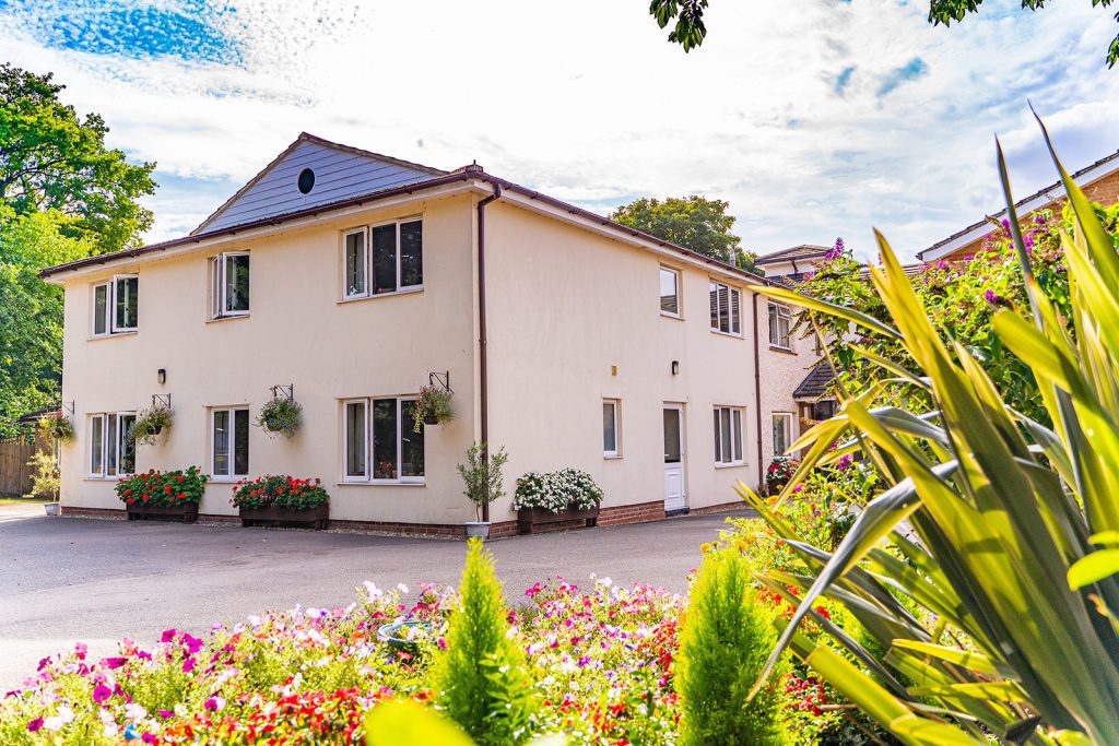 A light-coloured care home surrounded by vibrant flower beds and greenery, featuring large windows and a welcoming entrance.