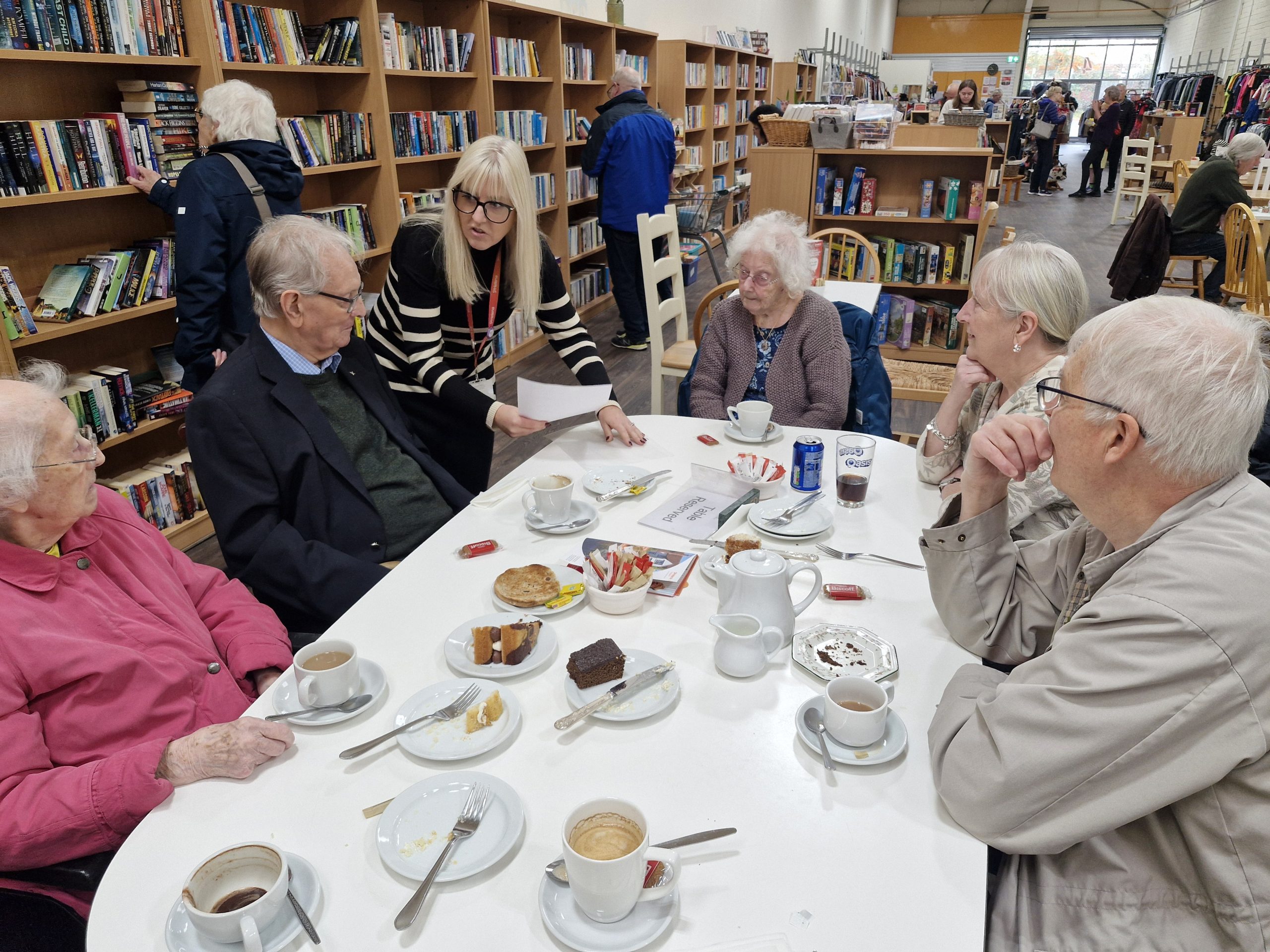 A group of six elderly people at a table with empty tea cups and plates in a bookshop, engaged in conversation with a staff member.