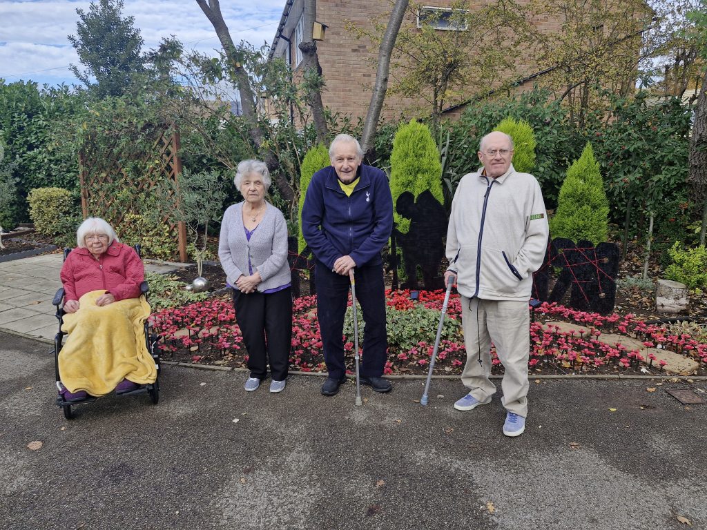 Four residents of Stanway Green Lodge stand together outside, surrounded by vibrant flowers and greenery, showcasing a warm community atmosphere.