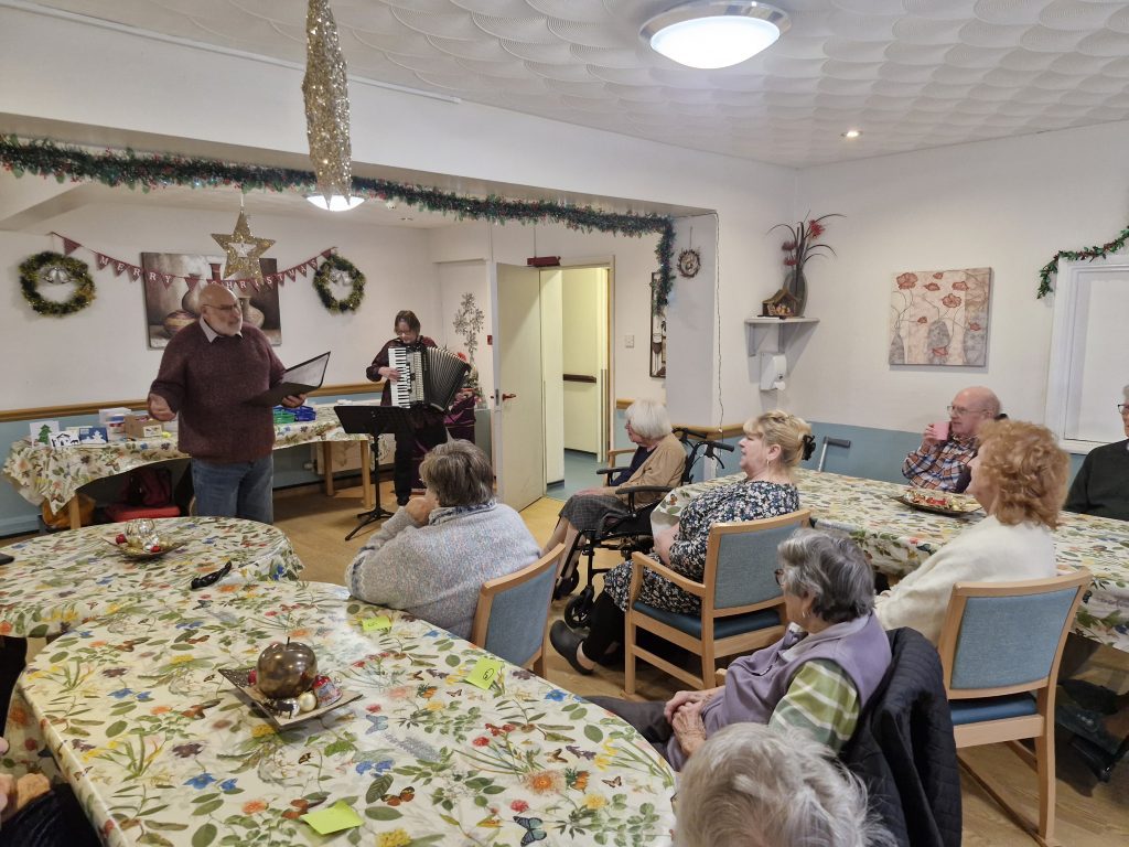 A festive activity in a warm, inviting lounge with residents seated around tables, enjoying live music from an accordion player.