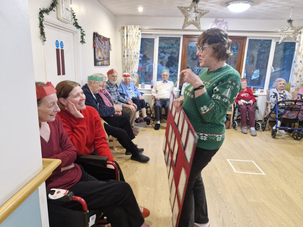 A festive gathering in a care home, with residents in Christmas hats smiling and a staff member engaging them, surrounded by decorations.
