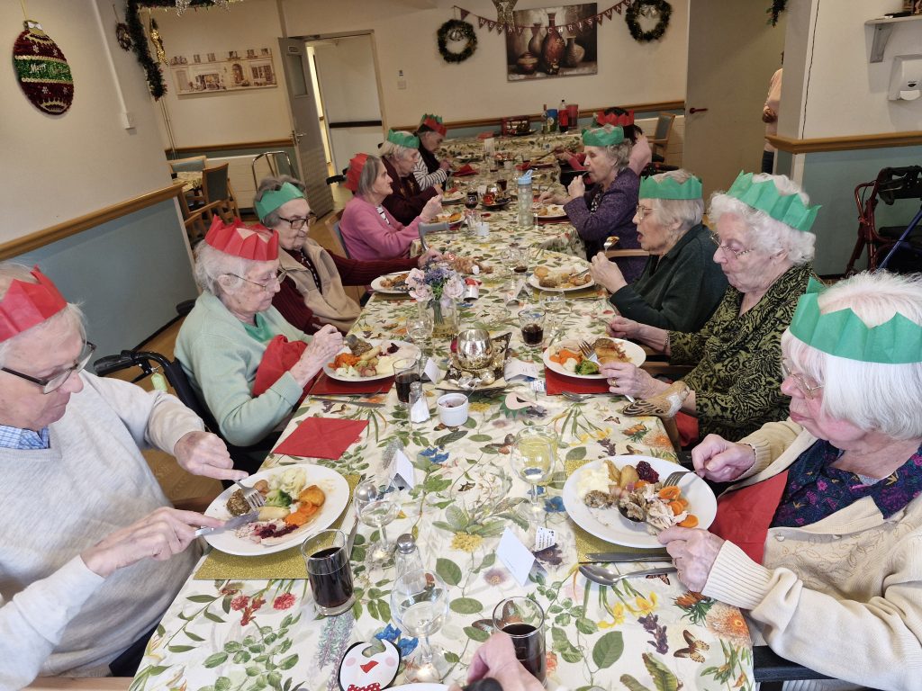 A festive dining scene with residents at long tables wearing paper crowns, enjoying a meal with colourful dishes and seasonal decorations.
