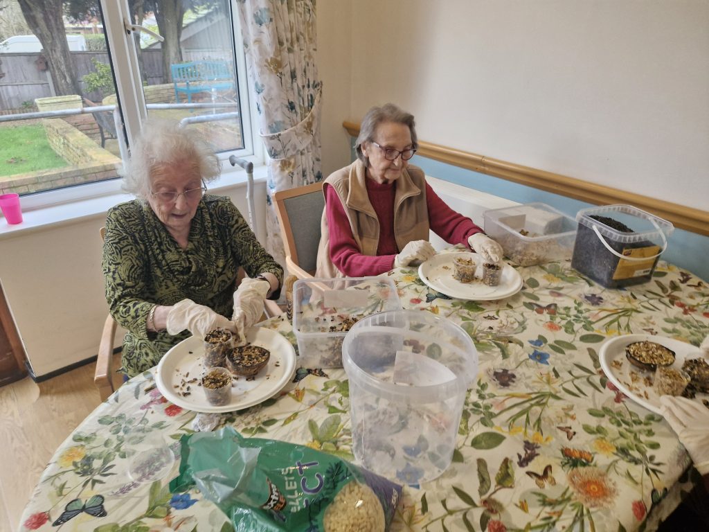 Two elderly women seated at a table, wearing gloves and crafting bird feeders from seeds, surrounded by materials and a bright, sunny room.