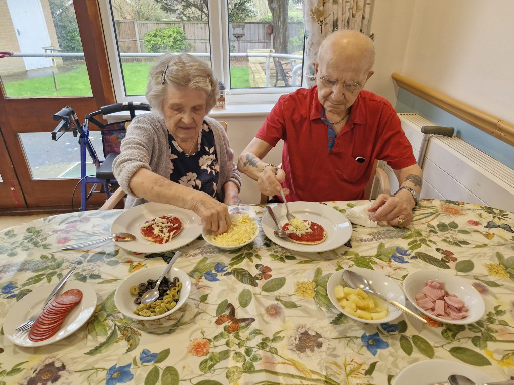 Two residents enjoying a meal at a table set with colourful dishes, including pasta and sliced meats, in a bright, homely dining area.