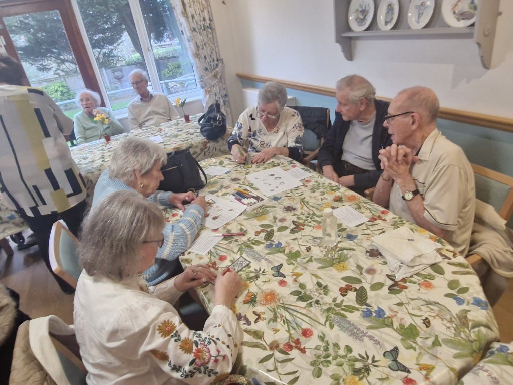Residents engage in activities around a floral-patterned tablecloth, smiling and interacting in a bright, welcoming room.