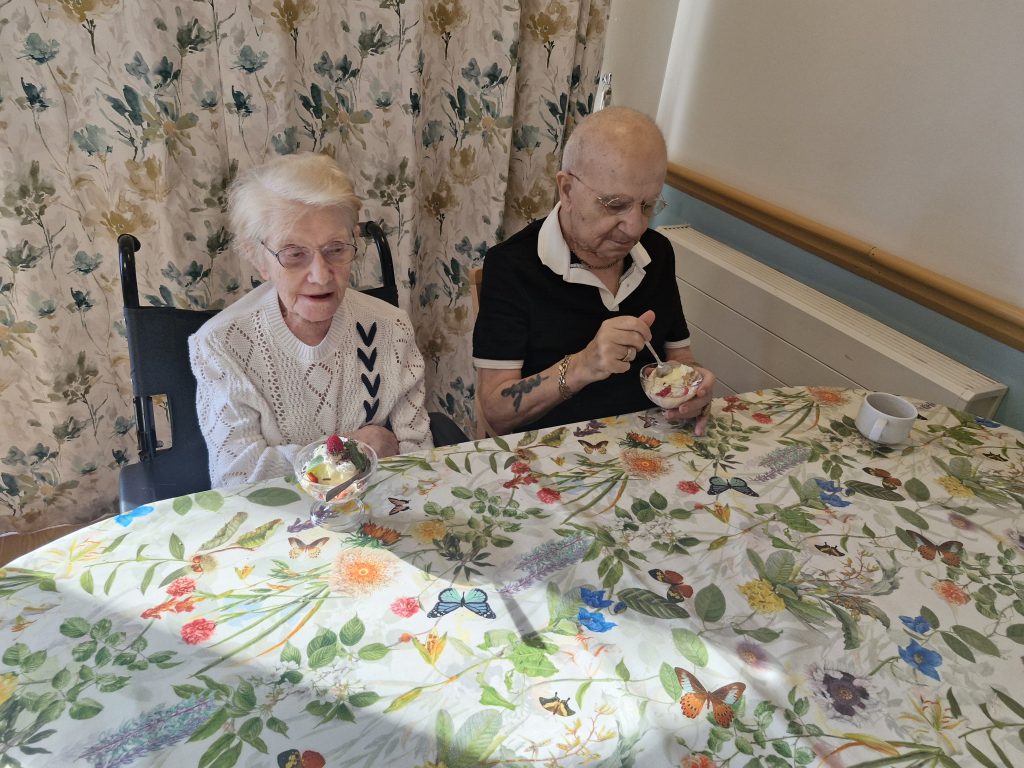 Two elderly women sit at a floral-patterned table, enjoying ice cream in bowls, with a bright and cheerful backdrop of curtains.