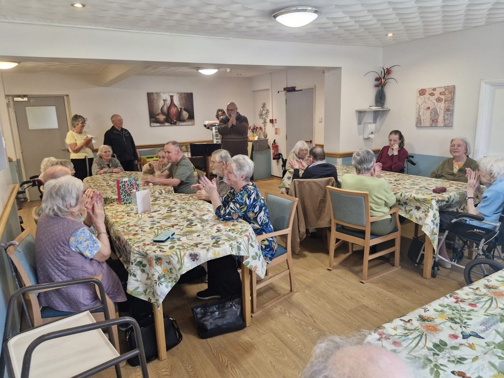 Residents and staff in a cheerful communal dining area, seated at tables adorned with floral tablecloths, engaging in lively conversation.