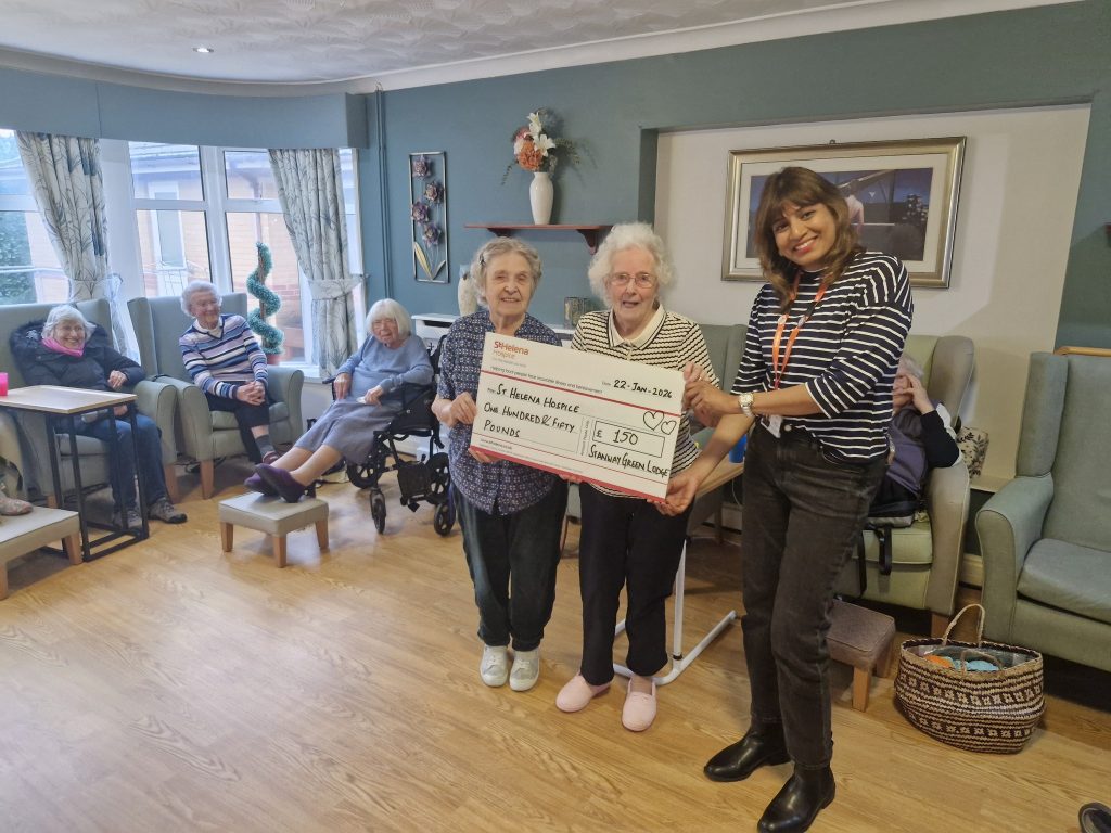 Two smiling elderly women hold a large cheque, while a staff member stands beside them. Residents in the background are seated in a cosy lounge setting.