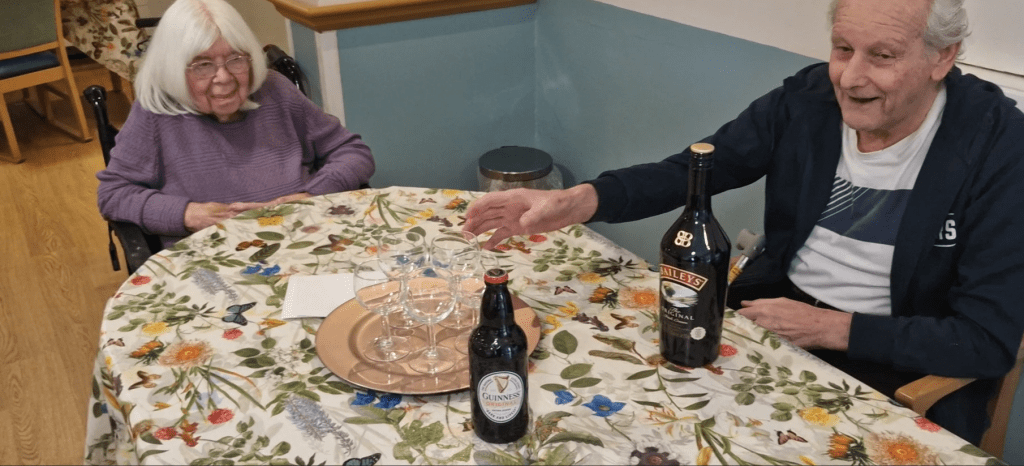 Two elderly residents sit at a floral tablecloth-covered table, smiling. There are glasses and bottles of drink, creating a warm, social atmosphere.